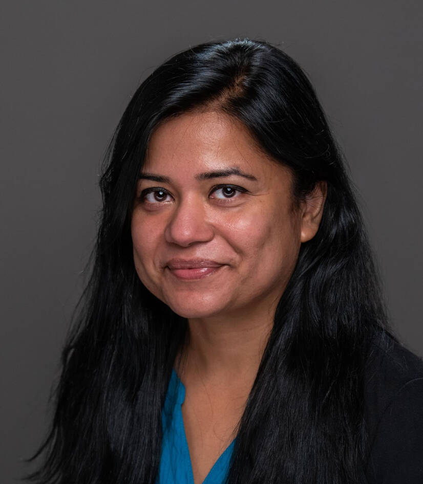 Naureen Hoque wearing a blue top, black jacket against a solid grey background.